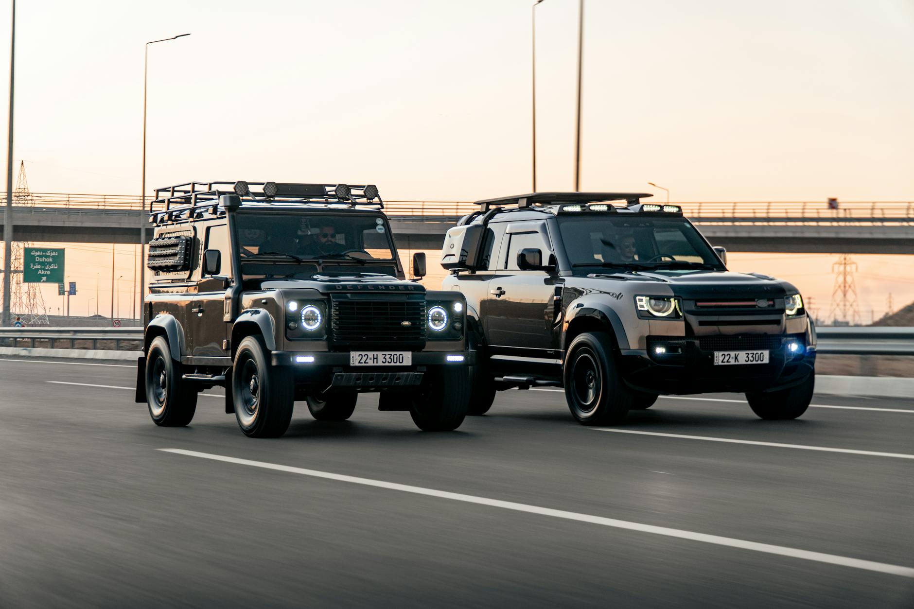 Dwa terenowe Land Rovery jadące obok siebie autostradą o zachodzie słońca